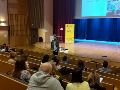 Mężczyzna stoi przy krawędzi sceny i przemawia do publiczności. Halowa sala z drewnianymi ławkami i kilkoma rzędami widzów patrzących w stronę sceny. Z tyłu sceny ciemnoniebieska kotara i żółty stojak informacyjny po prawej. Oświetlenie wewnętrzne, scena drewniana, widoczne rozrzucone kartki i sprzęt przy mówcy.