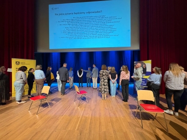 Około osiemnastu osób stoi na drewnianej scenie w sali konferencyjnej zwróconych twarzami do dużego, jasnego ekranu projekcyjnego. Scena ma niebieskie zasłony w tle i czerwone teatralne kotary po bokach. Po lewej i prawej stoją żółte słupki reklamowe. Na scenie stoją rozrzucone krzesła z czerwonymi siedziskami.