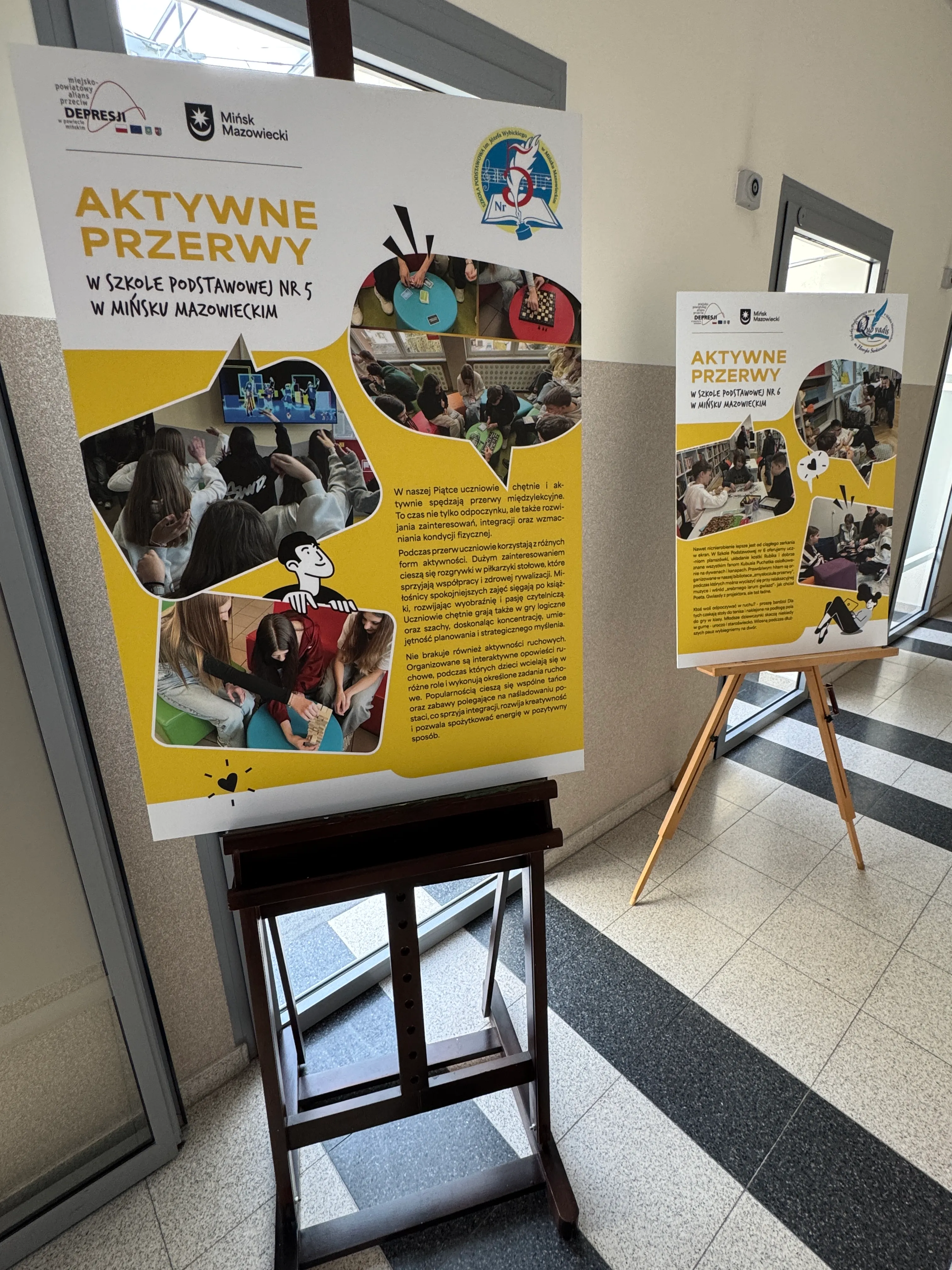 Dwa stojące plakaty o żółtych akcentach na drewnianych sztalugach ustawione w jasnym szkolnym korytarzu. Plakaty zawierają kilka owalnych zdjęć uczniów podczas zajęć i bloki tekstu; w tle widać drzwi z przeszkleniem i marmurową podłogę z ciemnymi pasami.