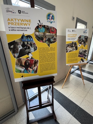 Dwa stojące plakaty o żółtych akcentach na drewnianych sztalugach ustawione w jasnym szkolnym korytarzu. Plakaty zawierają kilka owalnych zdjęć uczniów podczas zajęć i bloki tekstu; w tle widać drzwi z przeszkleniem i marmurową podłogę z ciemnymi pasami.