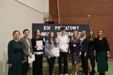 Grupa dziewięciu osób stoi na sali gimnastycznej przed tablicą z napisem dotyczącym konkursu ortograficznego. Trzy osoby w środku trzymają dyplomy. Tło stanowi czerwona ceglana ściana z siatką ochronną.