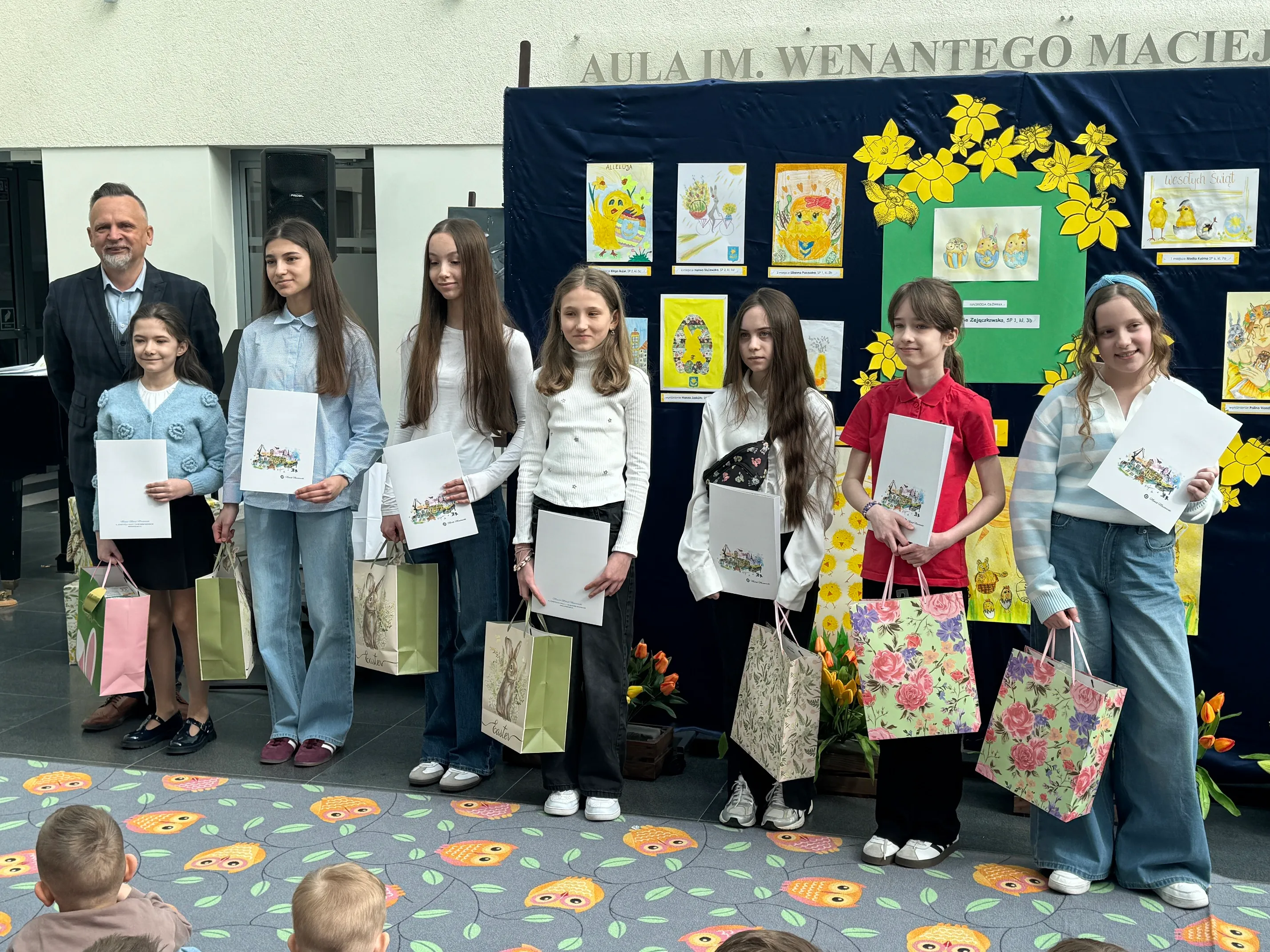 Siedem dziewczynek i dorosły mężczyzna stoją w rzędzie wewnątrz jasnego wnętrza. Dzieci trzymają białe teczki i kolorowe torby prezentowe. Za nimi stoi ciemnoniebieska tablica z kolorowymi rysunkami i żółtymi dekoracjami.