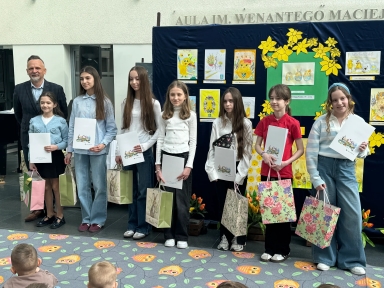 Siedem dziewczynek i dorosły mężczyzna stoją w rzędzie wewnątrz jasnego wnętrza. Dzieci trzymają białe teczki i kolorowe torby prezentowe. Za nimi stoi ciemnoniebieska tablica z kolorowymi rysunkami i żółtymi dekoracjami.