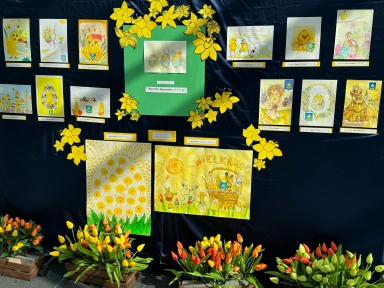 Kilkanaście wielkanocnych rysunków i pocztówek ułożonych na ciemnym tle.  
Dwa duże centralne plakaty w żółto-zielonej kolorystyce z napisem WIELKANOC.  
Żółte wycinanki kwiatów (żonkile) rozmieszczone wokół centralnej części.  
Kilka drewnianych skrzynek z żółto-pomarańczowo-czerwonymi tulipanami u dołu.