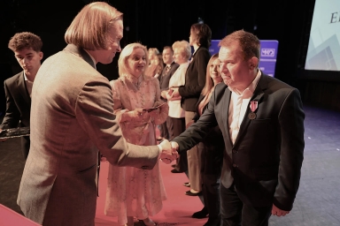 Dwaj mężczyźni ściskają sobie ręce na scenie podczas uroczystości. Mężczyzna po prawej ma medal na marynarce. W tle stoi kilku uśmiechniętych ludzi ustawionych w rzędzie. Otoczenie jest ciemne, scena oświetlona.