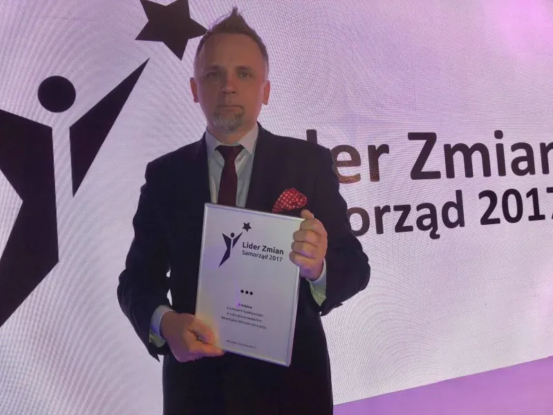 Mężczyzna w garniturze trzymający dyplom z napisem „Lider Zmian Samorząd 2017” na tle dużego wyświetlacza o podobnej treści.