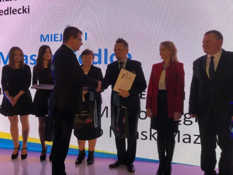 Grupa sześciu osób ubranych formalnie stoi na scenie, jedna z nich wręcza dokument drugiej podczas uroczystości.