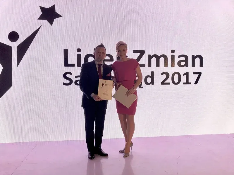 Mężczyzna w garniturze i kobieta w różowej sukience stoją na scenie z dokumentami na tle napisu „Lider zmian Samorząd 2017”.
