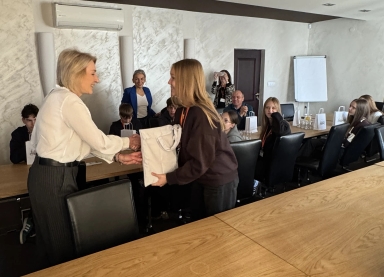 Kobieta wręcza upominek młodej osobie w sali konferencyjnej, wokół stołu siedzi grupa ludzi obserwujących tę scenę, jedna osoba robi zdjęcie telefonem, na stole stoją białe torby i szklanki z napojami.