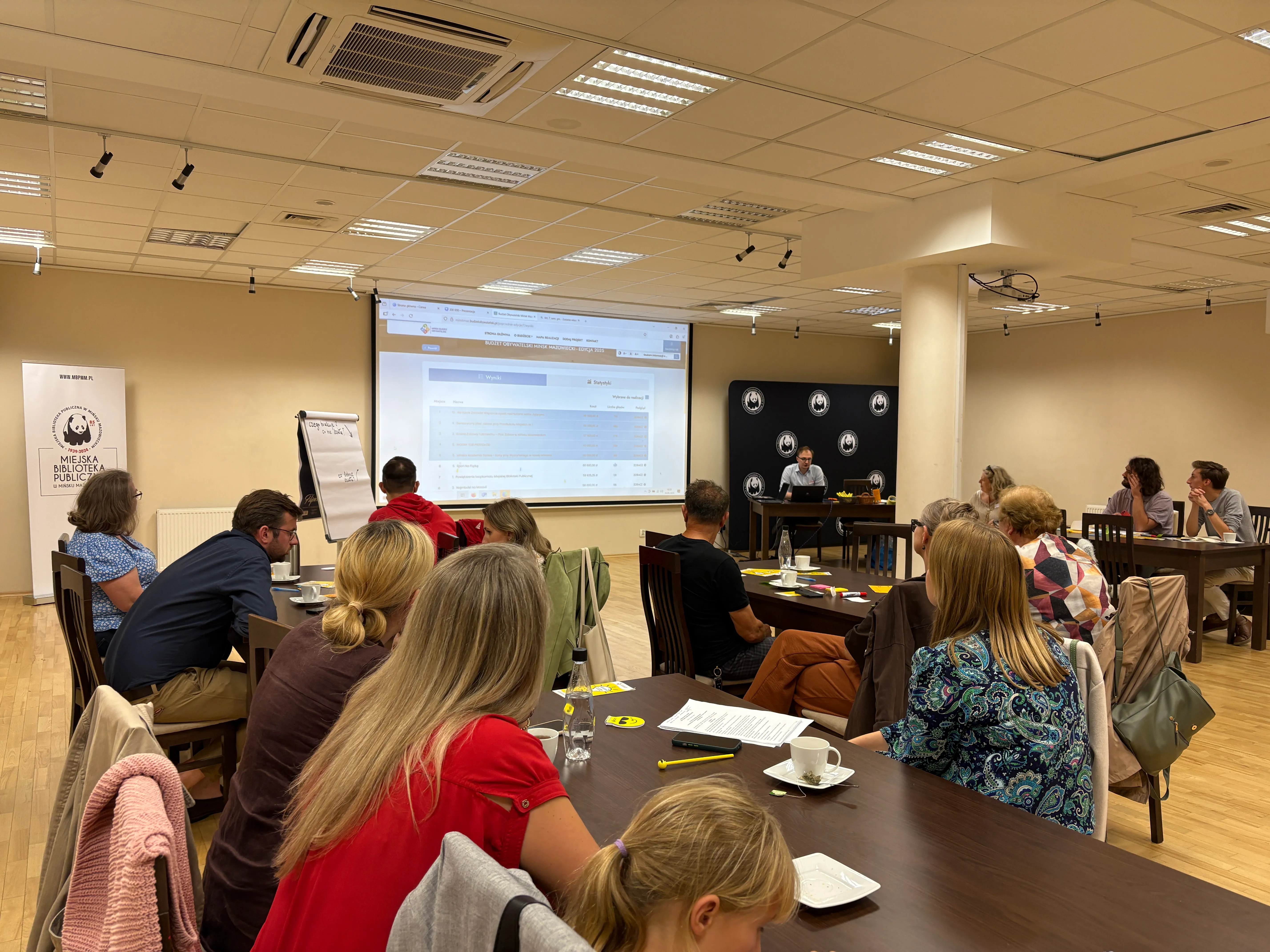 Grupa osób siedzi przy stołach w sali konferencyjnej, słuchając prezentera przy projektorze wyświetlającym stronę internetową, w tle widoczny roll-up z napisem miejskiej biblioteki publicznej.