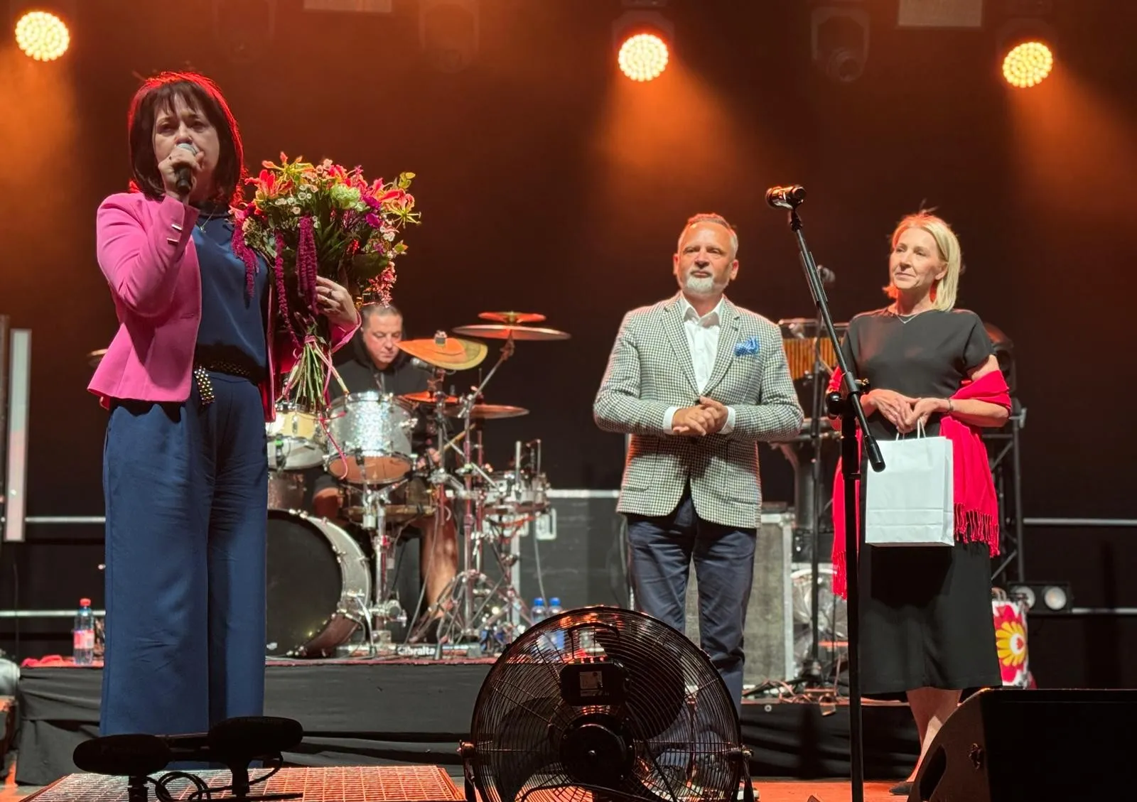 Trzy osoby na scenie podczas wydarzenia – kobieta z lewej mówi do mikrofonu i trzyma bukiet kwiatów, mężczyzna i kobieta z prawej stoją blisko mikrofonu, w tle perkusista na instrumentach.
