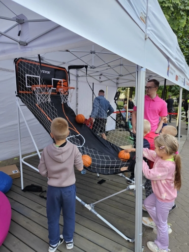 Dzieci grające w koszykówkę na ulicznym stoisku pod zadaszeniem, obok stoi mężczyzna w różowej koszulce.