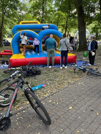 Dzieci bawią się na niebiesko-żółto-czerwonej dmuchańcej zjeżdżalni w parku, obok stoją osoby dorosłe, a na pierwszym planie leżą rowery i hulajnoga.