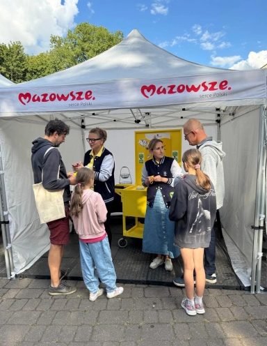 Pod namiotem z napisem „Mazowsze serce Polski” stoi pięć osób: dwie kobiety rozmawiające z mężczyzną i dwiema dziewczynkami na zewnątrz w dzień.