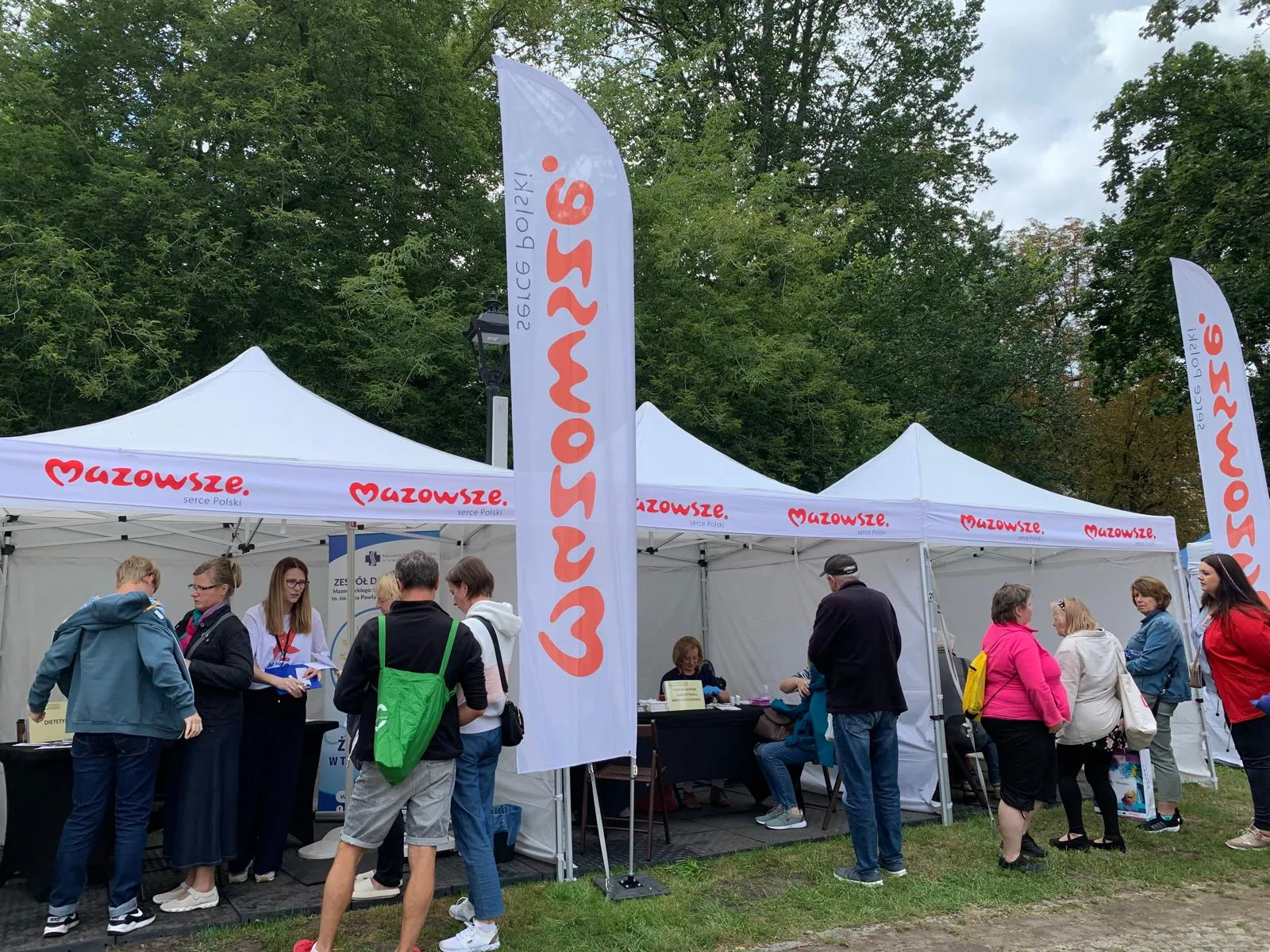 Na zewnątrz stoi kilka białych namiotów eventowych z napisem Mazowsze, przed nimi kilkanaście osób uczestniczy w wydarzeniu pod drzewami w ciągu dnia.