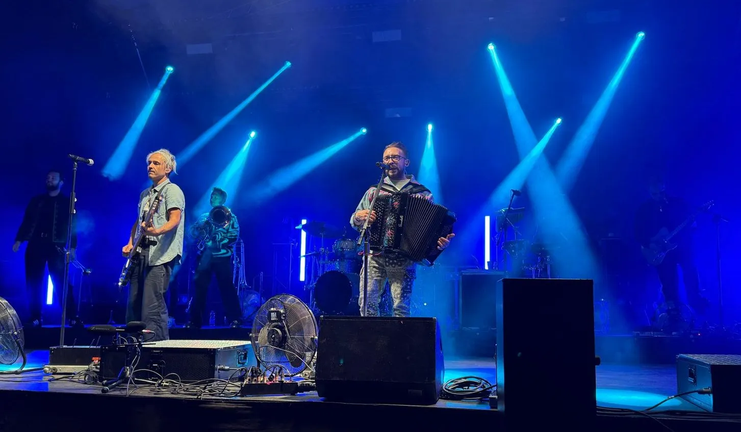 Zespół gra na scenie podczas koncertu z efektem niebieskiego oświetlenia i dymem, na pierwszym planie dwóch muzyków, jeden z gitarą elektryczną, drugi z akordeonem.