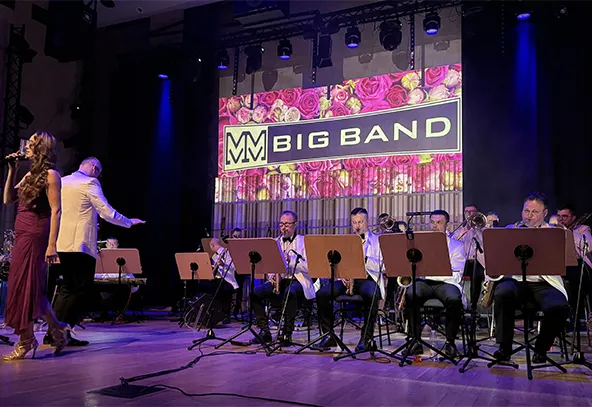 Na scenie sala koncertowa. Z przodu siedzi sekcja dęta w białych marynarkach przy pulpitach muzycznych. Po lewej stoi kobieta w długiej sukni i śpiewa, przed nią stoi dyrygent zwrócony do muzyków. W tle duży, podświetlony panel z kwiatowym motywem. Scena oświetlona fioletowo-niebieskim światłem.