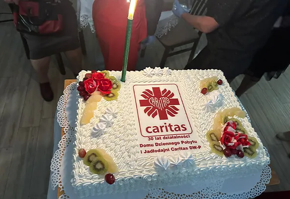 Prostokątny biały tort z bitą śmietaną, udekorowany plasterkami kiwi, czerwonymi owocami i czerwonymi kwiatami z lukru, z zieloną świecą. Na torcie nadrukowane logo i napis Caritas oraz informacja o 30 latach działalności. Tort stoi na białej serwetce na stole, w tle ludzie stojący w pomieszczeniu.