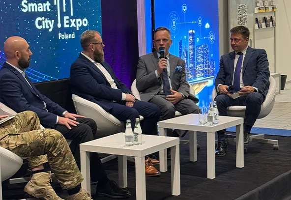 Czterech mężczyzn siedzi na białych fotelach wokół dwóch białych stolików na scenie z czarną podłogą. Jeden mężczyzna w garniturze mówi do mikrofonu. Tło z niebieskimi grafikami i napisem „Smart City Expo Poland”.