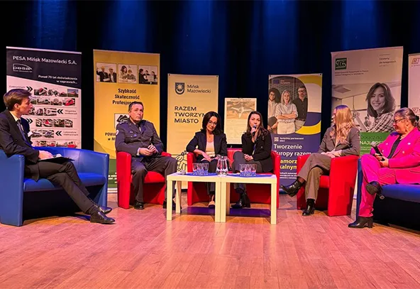 Sześć osób siedzi na scenie w czerwonych i niebieskich fotelach. Scena jest wewnątrz, ma drewnianą podłogę i ciemnoniebieskie tło. Za uczestnikami stoją pionowe banery. Na środku stoi niski biały stolik z dzbankiem wody i szklankami. Jedna kobieta trzyma mikrofon i mówi.