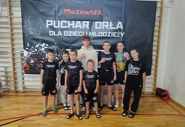 Grupa ośmiu dzieci i młodzieży stoi na sali gimnastycznej przed czarnym banerem z napisem „Mazowsze. Puchar Orła dla dzieci i młodzieży”. Ubrani są głównie w czarne sportowe stroje. W tle widać drewniane drabinki ścienne.