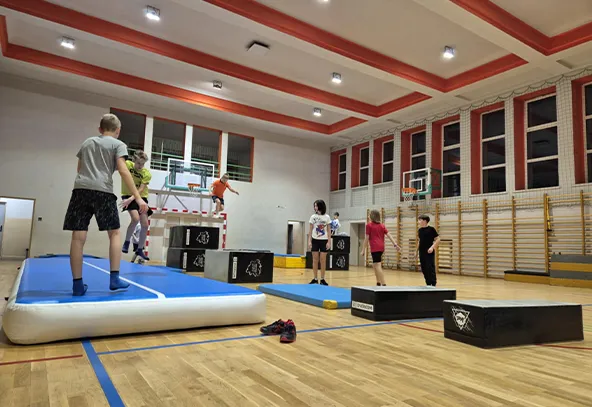Wewnątrz hali sportowej grupa dzieci ćwiczy na torze przeszkód z matami i skrzyniami gimnastycznymi. Pomieszczenie ma jasne ściany, czerwone akcenty na suficie i wysokie okna. Na podłodze leżą buty.