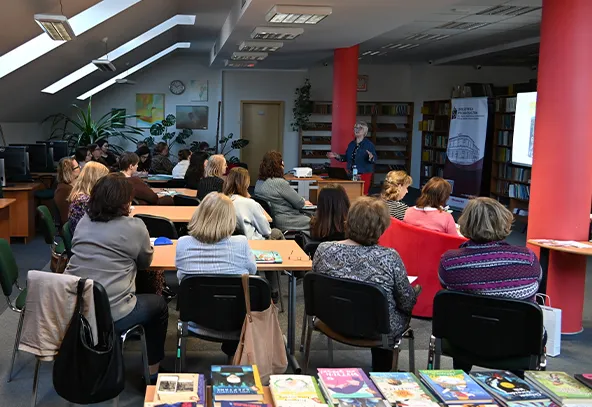 Grupa około dwudziestu osób siedzi przy stolikach w czytelni lub sali szkoleniowej. Prowadząca stoi przy ekranie i projektorze z przodu pomieszczenia. Na pierwszym planie leżą kolorowe książki, w tle regały, czerwone słupy i okna dachowe wpuszczające światło.