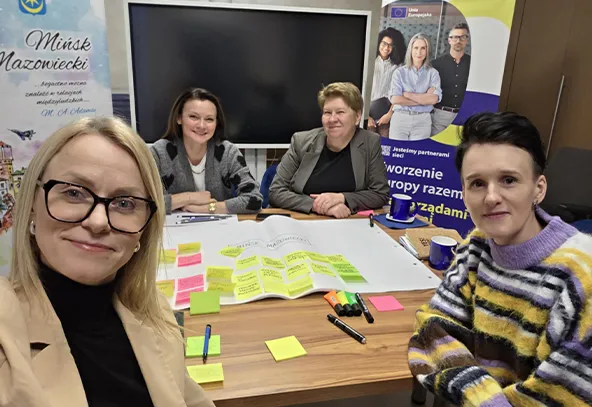 Cztery kobiety siedzą przy stole wewnątrz pomieszczenia. Kobieta z przodu po lewej robi selfie. Na stole leży duży arkusz z żółtymi, zielonymi i różowymi karteczkami samoprzylepnymi, markerami i kubkami. W tle widać duży ekran i stojące banery.