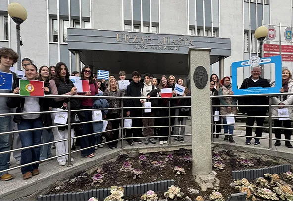 Grupa około 25 osób stoi przed wejściem do budynku urzędu miasta. Ludzie stoją przy metalowej balustradzie i trzymają małe flagi różnych krajów i kartki. Jest dzień, na pierwszym planie znajduje się rabatka z ozdobną kapustą. Budynek ma wielopiętrową fasadę i wejście z daszkiem.