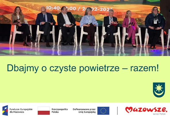 Siedzi siedem osób w rzędzie na białych plastikowych krzesłach na scenie. Tło sceny ma kolorowe, pomarańczowo-zielono-żółte wzory i odbija się na podłodze. Przed sceną znajduje się jasny zielonkawy pas z kilkoma logotypami u dołu.