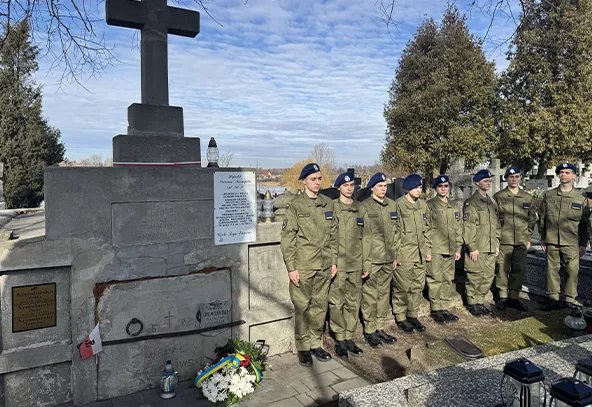 Grupa dziewięciu umundurowanych żołnierzy stoi w równym szeregu przy kamiennym grobowcu z dużym krzyżem. Na ziemi obok nagrobka leży biało-niebieski wieniec i zapalone znicze. Tło stanowią drzewa i jasno-niebieskie niebo w ciągu dnia.