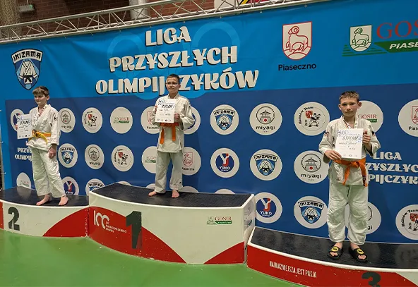 Trzech chłopców w białych judogach z pomarańczowymi pasami stoi na trzystopniowym podium w hali sportowej. Każdy trzyma dyplom. Za nimi niebieskie tło z okrągłymi emblematami i logotypami. Zielona podłoga.