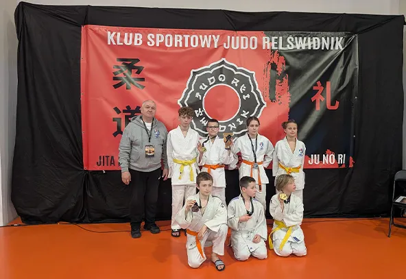 Osiem osób stoi i klęczy na pomarańczowej macie w sali. Siedmioro dzieci nosi białe judogi z żółtymi i pomarańczowymi pasami. Cztery dzieci stoją w drugim rzędzie, trzy klęczą z przodu i trzymają medale. Po lewej stoi dorosły trener w szarej bluzie. W tle duży czerwono-czarny baner z okrągłym logo klubu.