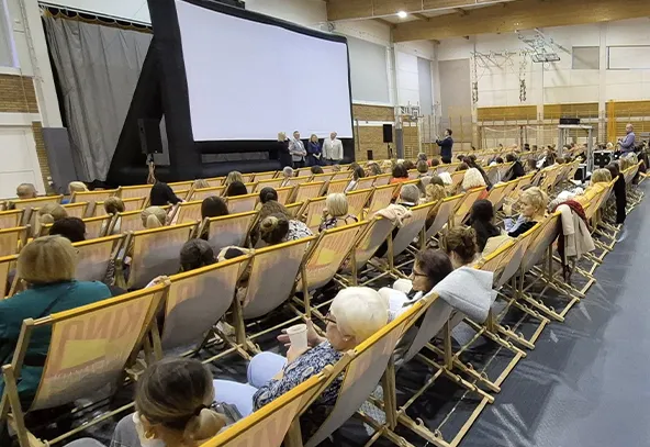 Wewnątrz dużej hali sportowej ustawione są rzędy leżaków, na których siedzi wiele osób zwróconych w stronę dużego, białego ekranu. Przed ekranem stoi trzech mężczyzn, a obok nich stoi osoba z mikrofonem. Tło hali jest jasne, z widocznymi elementami konstrukcyjnymi i sprzętem sportowym.