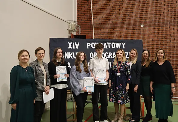 Dziewięć osób stoi na hali sportowej na tle planszy z napisem dotyczącym konkursu ortograficznego. Troje młodych ludzi w środku trzyma dyplomy. Wszyscy patrzą w stronę aparatu.