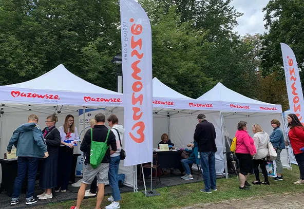 Grupa osób stoi przy białych namiotach z napisem Mazowsze na zewnątrz w zielonym parku w ciągu dnia.