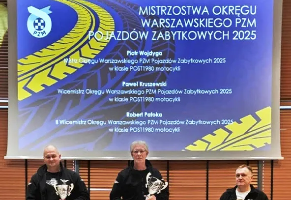 Trzej mężczyźni siedzą na scenie i każdy trzyma puchar. Za nimi wisi duży ekran z niebieskim tłem i żółtym motywem bieżnika opony. Scena znajduje się wewnątrz sali z drewnianymi panelami na ścianie. Mężczyźni mają ciemne ubrania; środkowy ma siwe włosy.