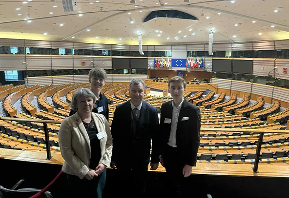 Cztery osoby stoją na balkonie dużej sali konferencyjnej z półkolistym układem rzędów siedzeń. W tle widoczne flagi i duży niebieski ekran z symbolem Unii Europejskiej. Sala jest dobrze oświetlona.