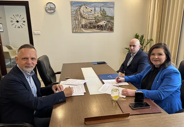 Trzy osoby siedzą przy długim drewnianym stole w biurze. Dwaj mężczyźni i kobieta zajmują miejsca po obu stronach stołu. Kobieta i mężczyzna po lewej podpisują dokumenty. Na stole leżą teczki, papiery, telefon i szklanka z napojem. W tle widoczny obraz na ścianie, zegar i roślina doniczkowa.