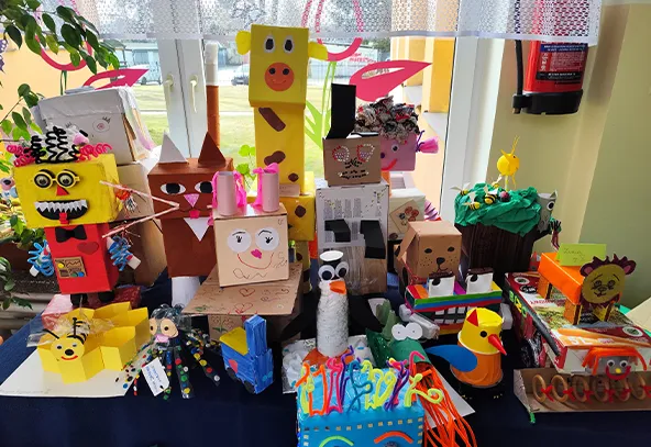 Stołowa wystawa kolorowych, ręcznie wykonanych pudełkowych zwierząt i postaci ustawionych przy oknie w świetle dziennym. Różnokolorowe figurki mają twarze, oczka, rogi i ozdoby z papieru, filcu i rurek, ułożone na granatowym obrusie. W tle widać firankę, zieleń roślin i gaśnicę na ścianie.