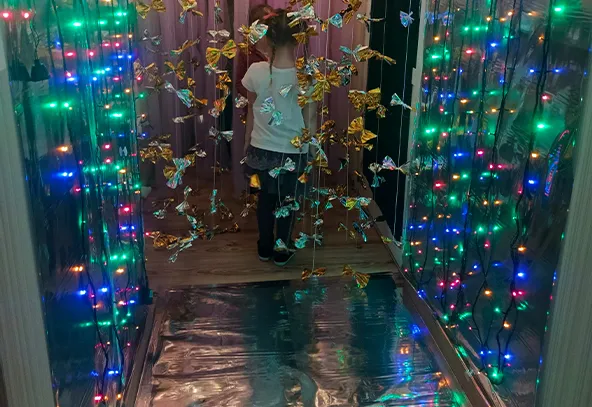 Dziecko stoi tyłem w wąskim, wewnętrznym korytarzu. Po bokach wiszą kolorowe, migające lampki. Przed dzieckiem zwisają złote i srebrne metaliczne dekoracje. Podłoga ma odblaskową folię odbijającą światła.