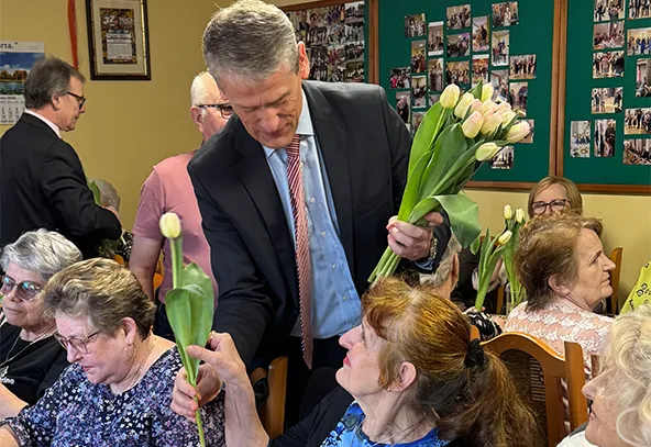 Mężczyzna w garniturze wręcza białe tulipany starszym kobietom siedzącym przy stołach w jasnej sali. W tle zielona tablica z wieloma fotografiami.