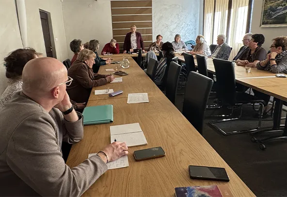 Grupa około trzynastu osób siedzi przy długim drewnianym stole w sali konferencyjnej. Kobieta stoi na końcu stołu i przemawia. Uczestnicy notują, obok leżą notesy i smartfony. Wnętrze jest jasne, po prawej okna z żaluzjami, ściany w jasnych kolorach.