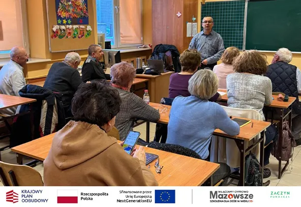 Grupa starszych osób siedzi przy stolikach w sali lekcyjnej, mężczyzna stoi przy tablicy i mówi do grupy. W tle okna i dekoracje ścienne.