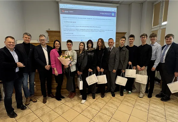 Trzynastu dorosłych i nastolatków stoi w rzędzie w sali konferencyjnej. Na ścianie za nimi widać rzutnik wyświetlający stronę internetową. Kilka osób trzyma białe torby upominkowe, jedna osoba trzyma bukiet kwiatów. Podłoga jest kafelkowana, pomieszczenie jest oświetlone wewnątrz.