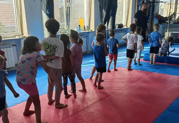 Dzieci ustawione w szeregu na sali gimnastycznej, ubrani w sportowe stroje, uczestniczą w zajęciach pod okiem instruktora.