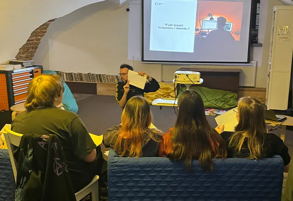 W sali warsztatowej pięć osób siedzi tyłem na kanapie i krzesłach, patrząc na prowadzącego siedzącego z przodu. Prowadzący pokazuje kartkę i mówi do grupy. Z przodu stoi projektor rzutujący slajd na biały ekran. W tle widoczna ściana z ceglaną arkadą i niski regał z płytami.