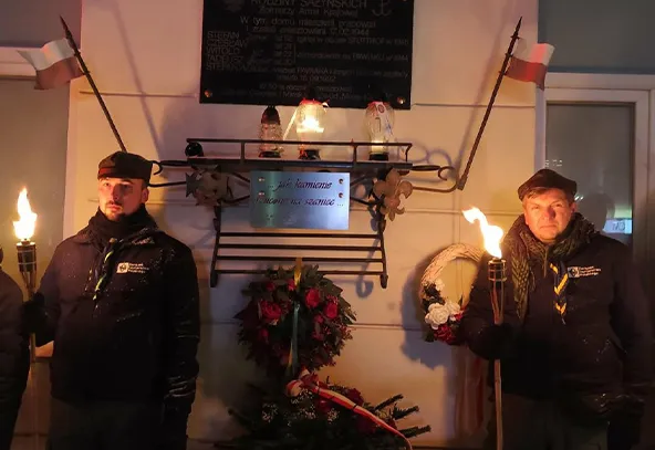 Dwaj mężczyźni stoją po obu stronach ściany, każdy trzyma palącą pochodnię. Są ubrani w ciemne kurtki i czapki. Na ścianie widoczna jest tablica pamiątkowa z wieńcem i kwiatami poniżej. Po bokach na drzewcach wiszą małe flagi Polski. Scena odbywa się nocą na zewnątrz.