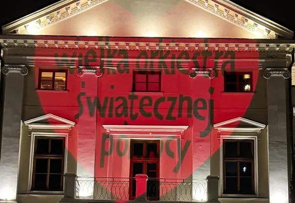 Budynek o klasycznej architekturze podświetlony na biało z czerwoną projekcją w kształcie serca i ciemnym napisem na froncie, widok nocny.