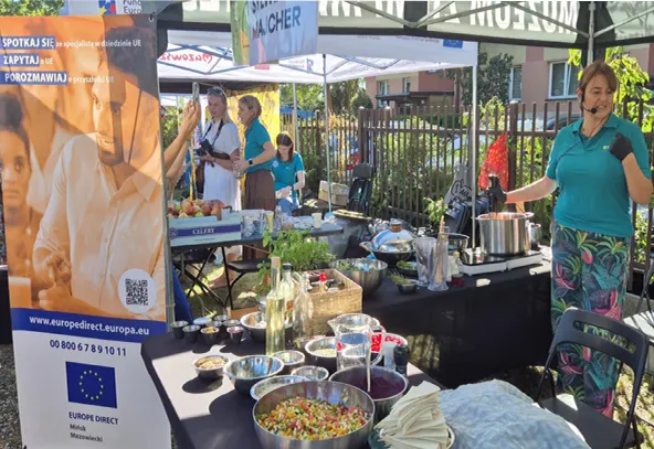 Stoisko na zewnątrz pod namiotem z kobietą na mikrofonie po prawej stronie, przy stole z różnymi miskami i butelkami oraz dużym garnkiem. Po lewej stronie baner z informacjami o UE. W tle trzy osoby rozmawiają przy innym stoisku. Za stoiskami widoczny płot i budynki. Słoneczny dzień.